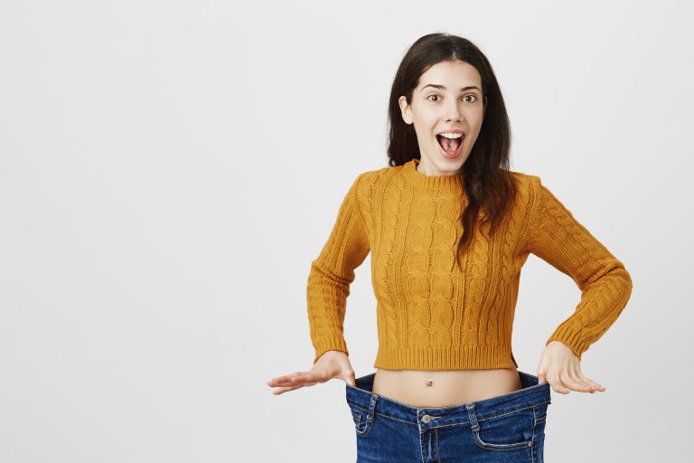 Astonished and happy young lady being impressed and excited because of losing weight, showing empty space in jeans by stretching it, standing over gray background. Girl is ready for summer weight loss