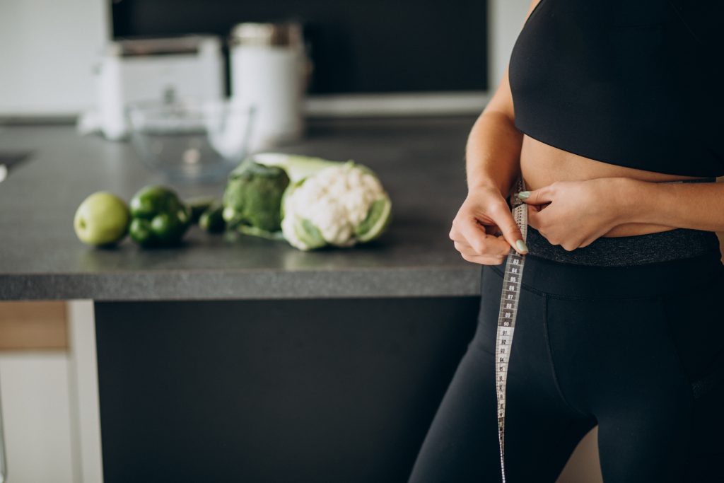 Astonished and happy young lady being impressed and excited because of losing weight, showing empty space in jeans by stretching it, standing over gray background. Girl is ready for summer weight loss